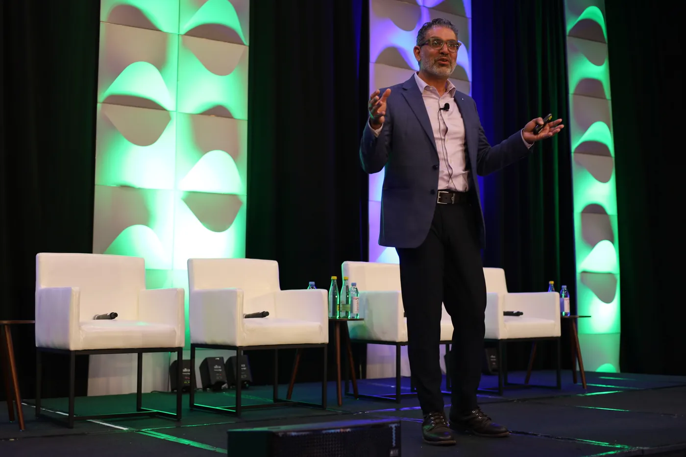 Moudy Elbayadi speaking on stage with green and blue lighting, gesturing mid-keynote